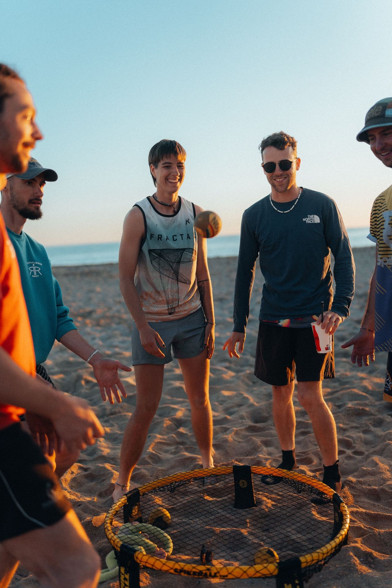 Group of people playing Spikeball on a sandy beach during sunset with the ocean in the background.