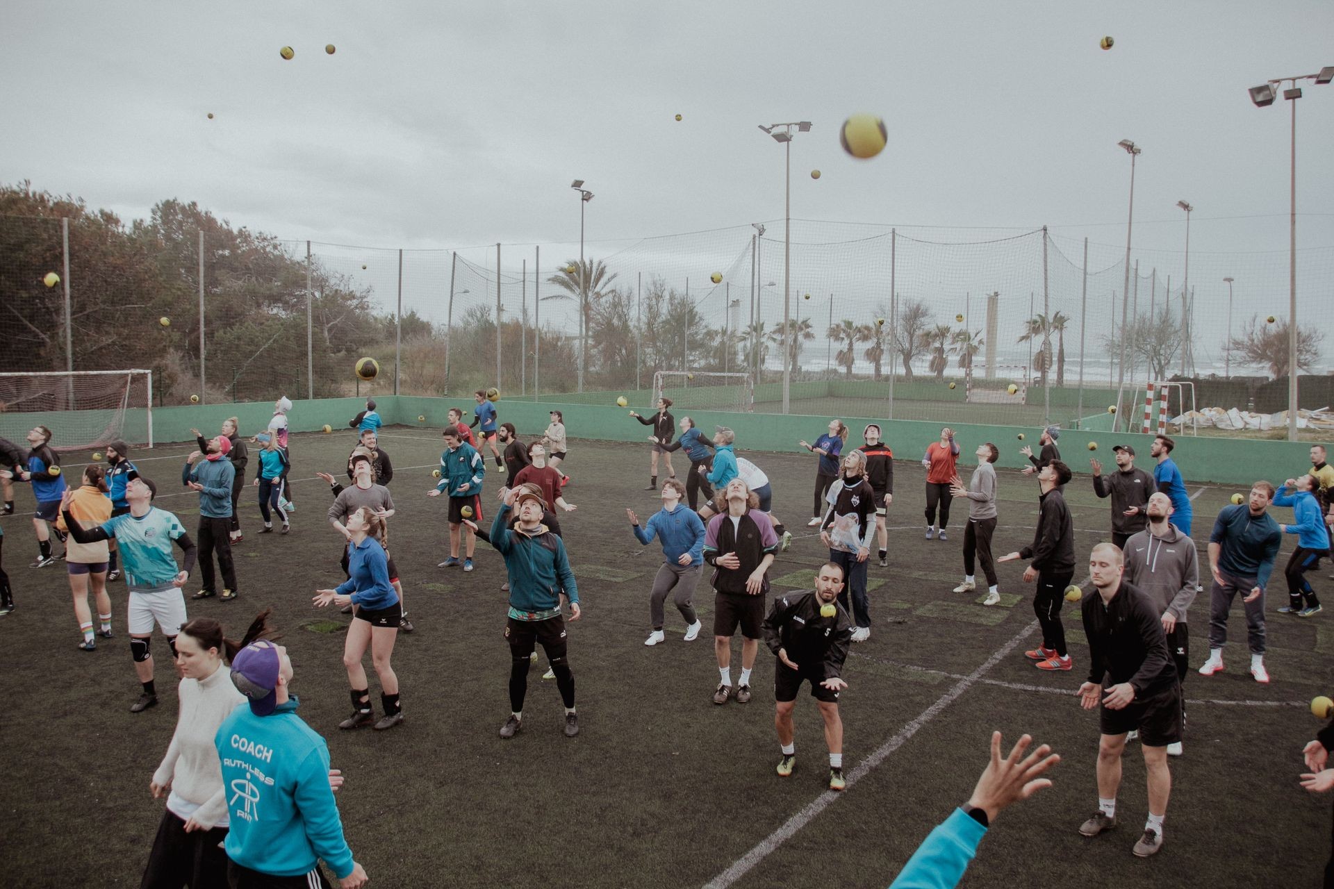 Group of people on an outdoor field simultaneously throwing and catching yellow balls in a coordinated exercise.