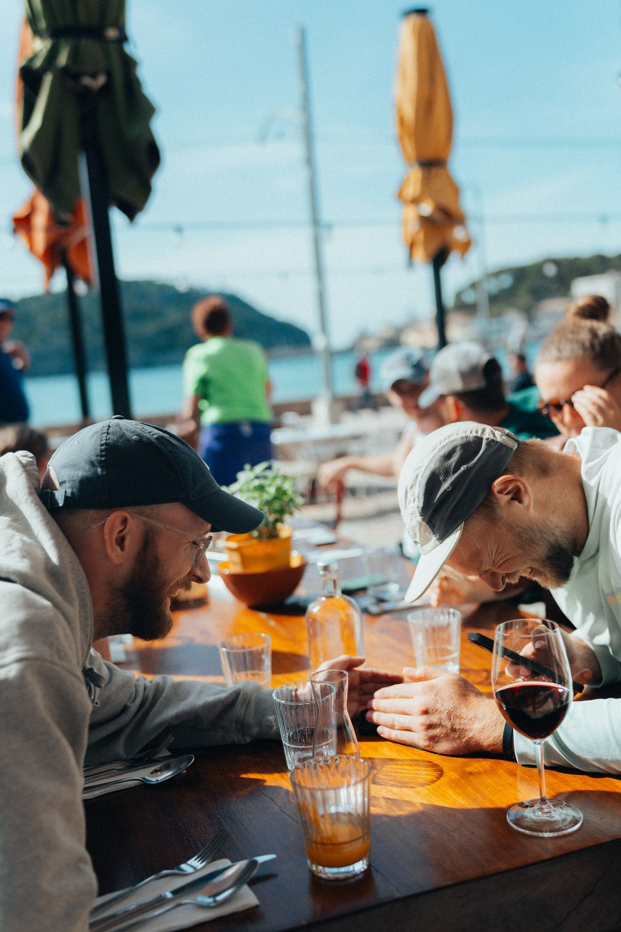 People enjoying drinks and conversation at an outdoor cafe by the water on a sunny day.