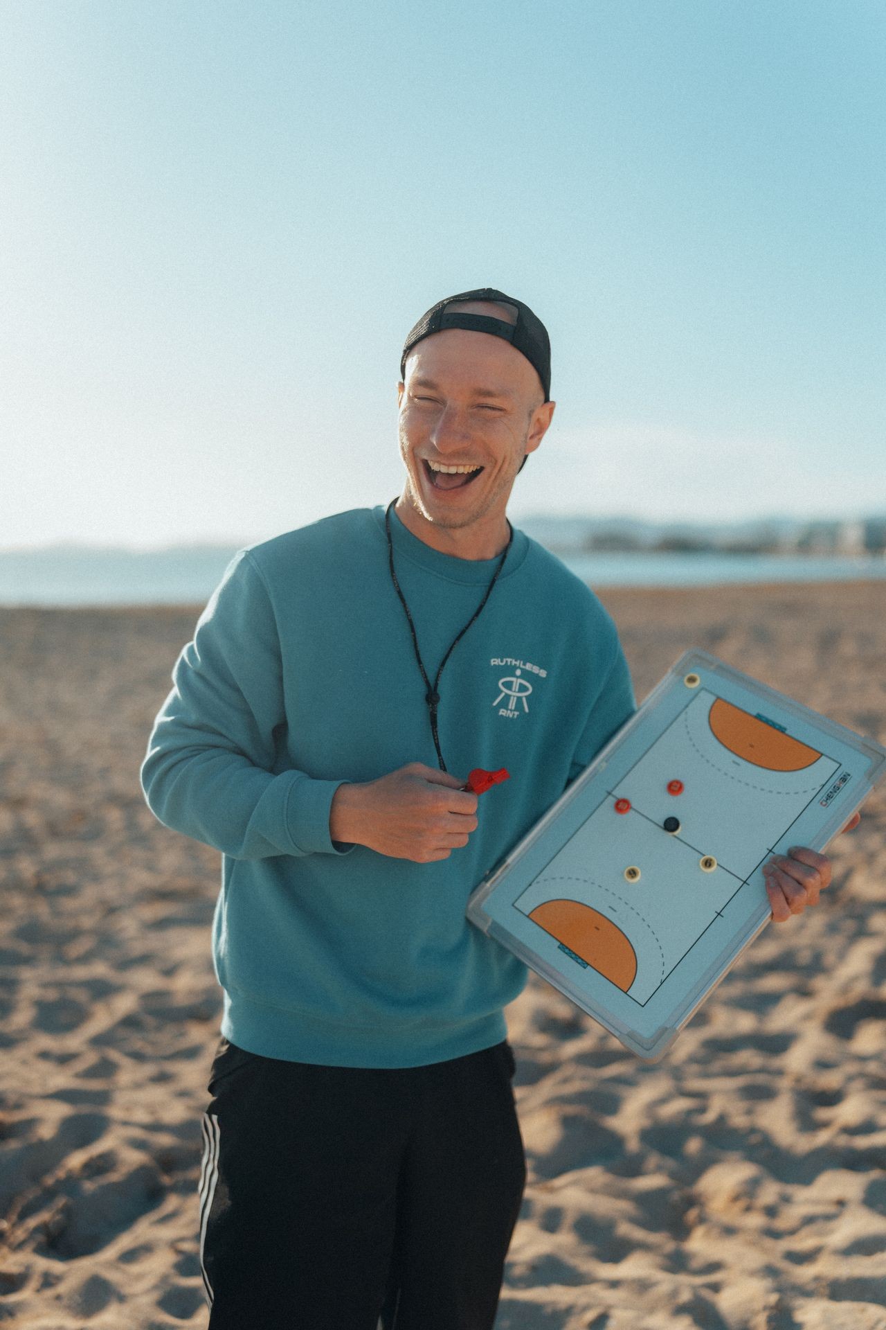 Man holding a strategy board on a sandy beach, laughing and wearing a cap and sweatshirt.