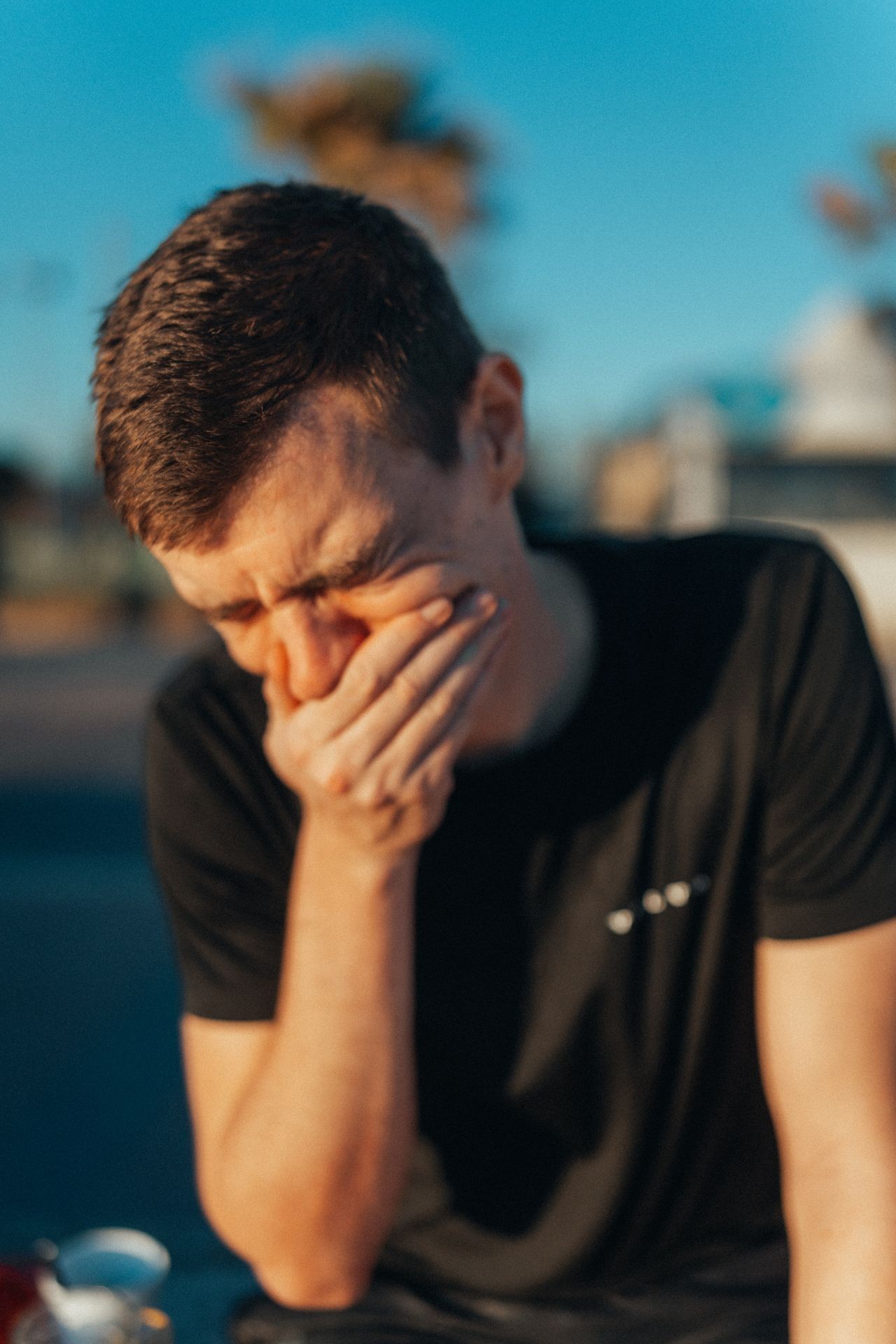 Person in a black t-shirt sitting outdoors with a blue sky and blurred trees in the background.